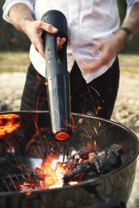 A person in a white shirt uses the Arteflame Outdoor Grills' Looft Lighter X: Revolutionary Charcoal Grill Lighter and Easy Wood Fire Starting to ignite a fire in a barbecue. The fire is producing small sparks and glowing coals, set against an outdoor backdrop with a grassy area.