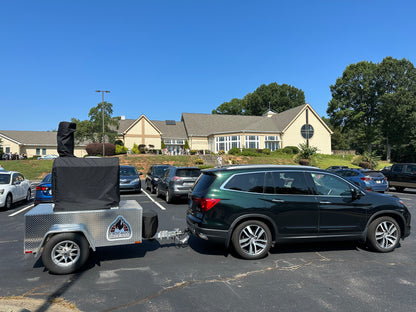 A green SUV is parked in a lot, towing a trailer labeled "Smoke Show." The trailer, equipped with covered equipment and featuring diamond plate detailing, prominently showcases the Chicago Brick Oven CBO 750 Tailgater. This hybrid pizza oven trailer has a 38" x 28" cooking surface, capable of baking 2-3 pizzas at a time or up to 50-60 pizzas an hour using both liquid propane and wood. In the background stands a large, light-colored building with a circular window and multiple dormers.
