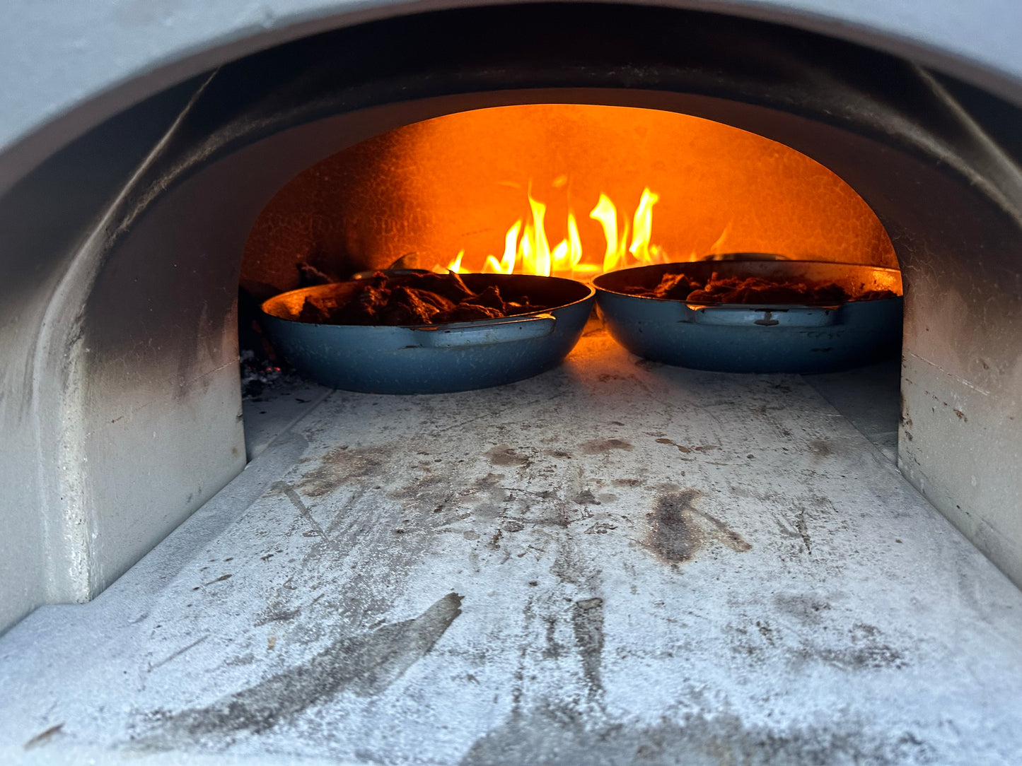 Close-up view of the Chicago Brick Oven CBO 750 Tailgater's interior, showing a blazing fire in the background. Two blue pans filled with brown, sizzling food are placed inside, capturing a rustic cooking scene. The hybrid pizza oven trailer's charred walls indicate frequent use, enhancing its authentic appeal.