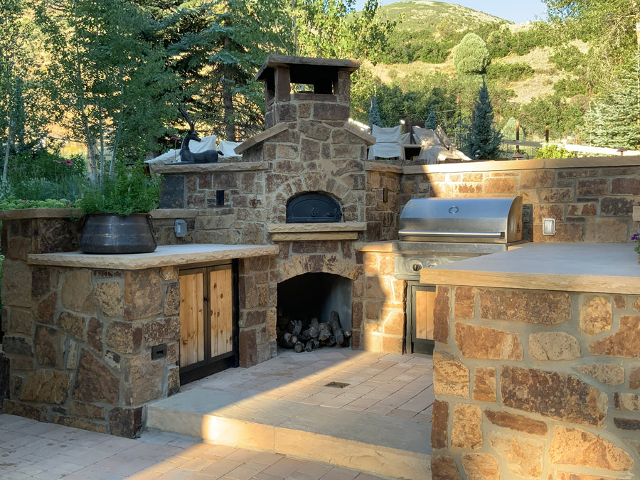 An outdoor kitchen with a stone backsplash featuring a wood-fired oven and a grill. The countertops are stone, and there are wooden cabinets underneath. The setting is surrounded by greenery and trees, with hills in the background. Firewood is stored below the oven.