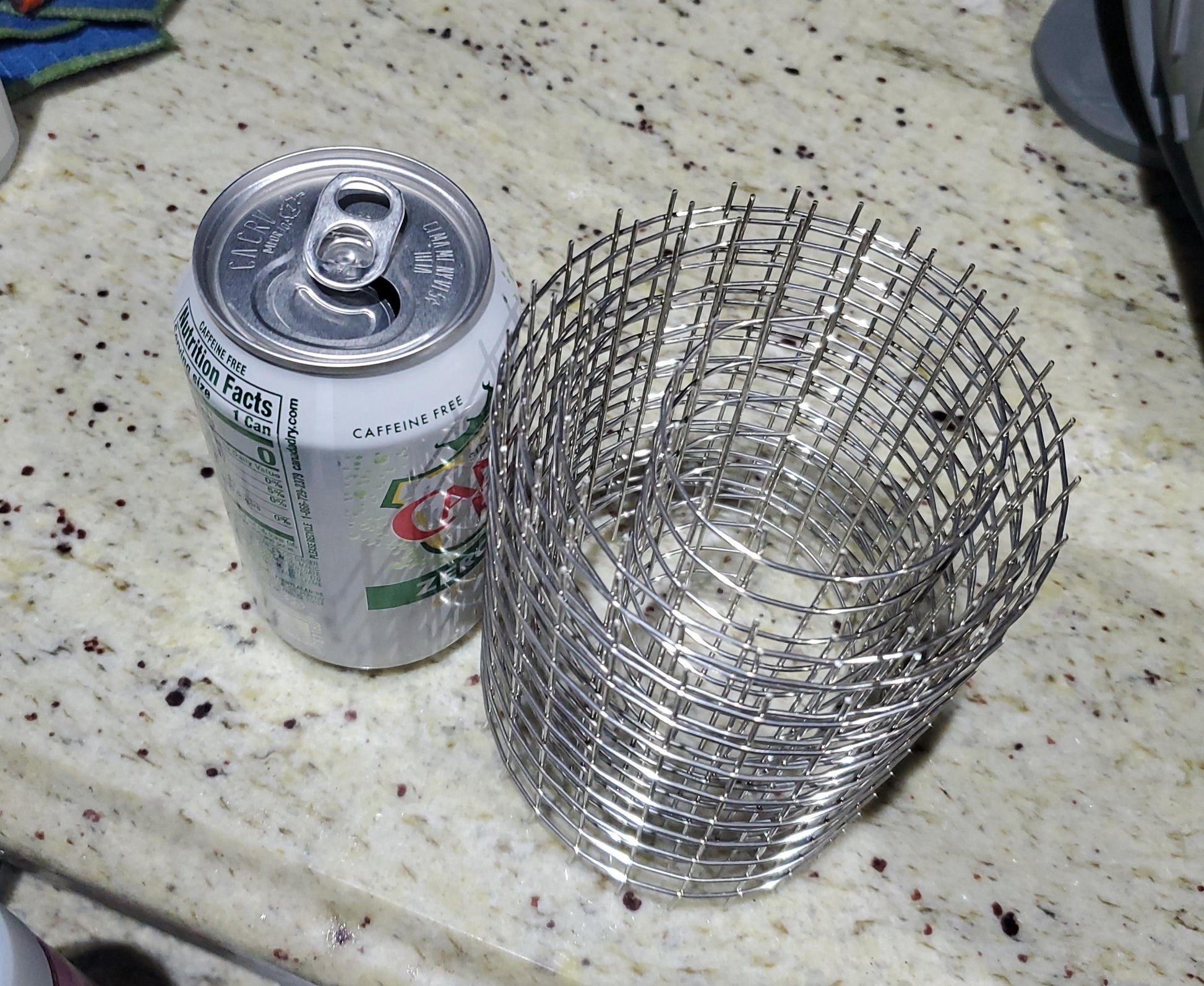 A can of Sprite Zero sits on a speckled countertop beside a cylindrical wire mesh organizer. The can is opened, and the wire mesh organizer, known as a DuraTech Spark Arrestor from Chicago Brick Oven, remains empty and stands upright like its stainless steel spark arrestor design.