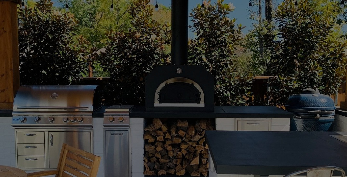 An outdoor kitchen features a variety of equipment, including a stainless steel gas grill, a pizza oven with wood storage below, and a ceramic smoker. The kitchen is set against a backdrop of lush greenery and trees. A wooden table is partially visible in the foreground.