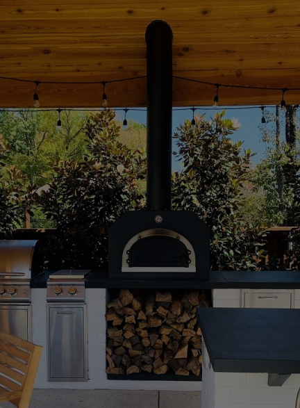 An outdoor kitchen with a built-in wood-fired pizza oven at the center, surrounded by storage cabinets and a grill on the left. The oven has a stack of firewood beneath it. The area is under a wooden pergola with string lights, and trees are visible in the background.