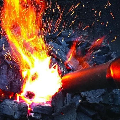 A roaring fire with bright orange flames and flying sparks, ignited from a pile of black charcoal. The Looft Lighter X: Revolutionary Charcoal Grill Lighter and Easy Wood Fire Starting by Arteflame Outdoor Grills, a cylindrical metal object that appears to be battery-powered, is pointed at the base of the fire, intensifying the flames. The background is dark and partially visible.