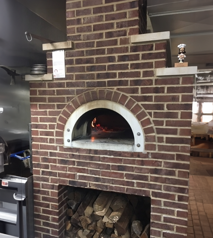 A brick pizza oven with a fire burning inside, set in a restaurant kitchen. Wooden logs are stored beneath the oven. A small chef figurine is placed on a ledge to the upper right of the oven. Cooking equipment is visible to the left.