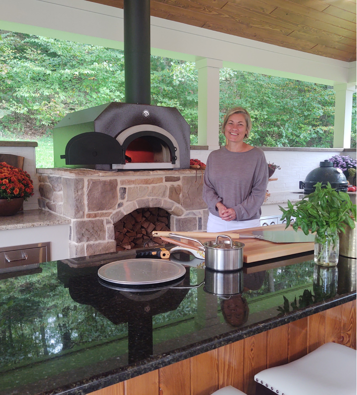 A woman stands smiling in an outdoor kitchen with a stone pizza oven behind her. The kitchen features a granite countertop, a cutting board with fresh basil, and various kitchen tools. There are trees in the background, creating a serene setting.