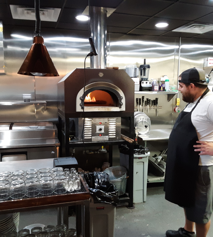 A man in a black apron and cap stands in a commercial kitchen, looking at a wood-fired pizza oven with a visible flame inside. The kitchen has stainless steel appliances, a prep area with glassware, and various utensils on the countertop.