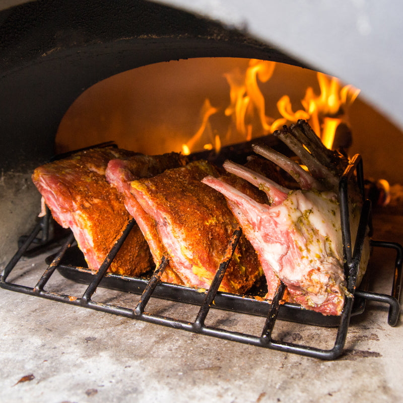 Inside the CBO 500 Countertop Wood Fired Pizza Oven by Chicago Brick Oven, seasoned cuts of meat rest on a roasting rack with flames flickering in the background. The upright-arranged meat racks display a beautifully crusted surface formed from an aromatic blend of spices and herbs.