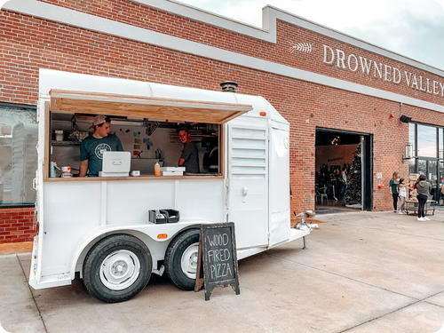 A white food truck serving wood-fired pizza is parked in front of a red brick building labeled "Drowned Valley." Two men are inside the truck, one wearing a cap. A small blackboard sign on the ground reads "Wood Fired Pizza." People are visible in the background.