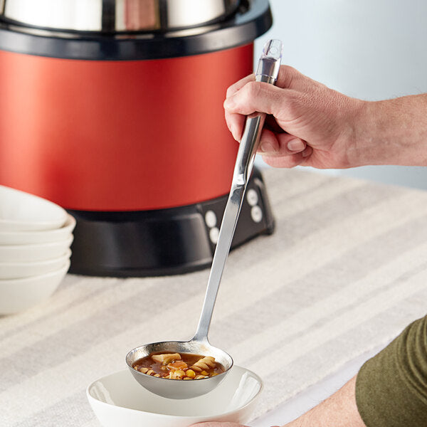 A person is using a Chicago Brick Oven 4 oz. Stainless Steel 12" Ladle to pour a serving of vegetable soup into a white bowl. Multiple empty bowls are stacked nearby on a striped tablecloth, and a red and black soup warmer is visible in the background.