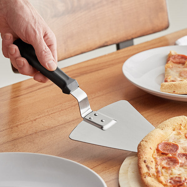 A hand holding a Chicago Brick Oven 13 1/4" Extra Large Pizza Pie Server with a black offset handle, lifting a slice of pepperoni pizza from a wooden pizza peel. Two white plates with slices of pizza are visible in the background, placed on a wooden table. This versatile serving tool from Chicago Brick Oven ensures every slice is served perfectly.