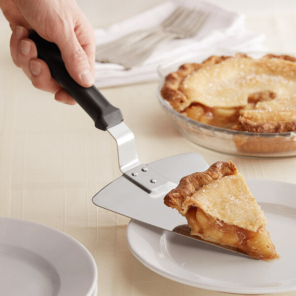 A hand using a Chicago Brick Oven 13 1/4" Extra Large Pizza Pie Server with Black Offset Handle to place a slice of pie onto a white plate. The pie has a golden-brown crust with a sugared top and is filled with apple filling. The remaining pie is in a glass dish in the background, resting on a beige tablecloth near some stacked plates and folded napkins.