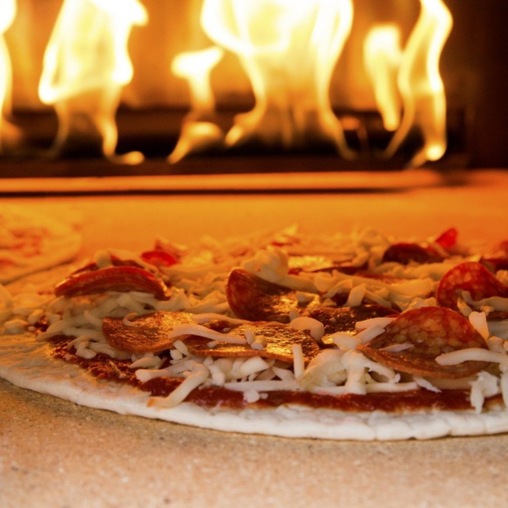 A close-up of a pizza topped with cheese and pepperoni, placed on a stone surface inside a Chicago Brick Oven CBO 750 Hybrid DIY Kit (Residential) | Dual-Fuel (Gas and Wood). Flames dance in the background, indicating that the pizza is baking perfectly in this outdoor oven lined with refractory cement for optimal heat retention.