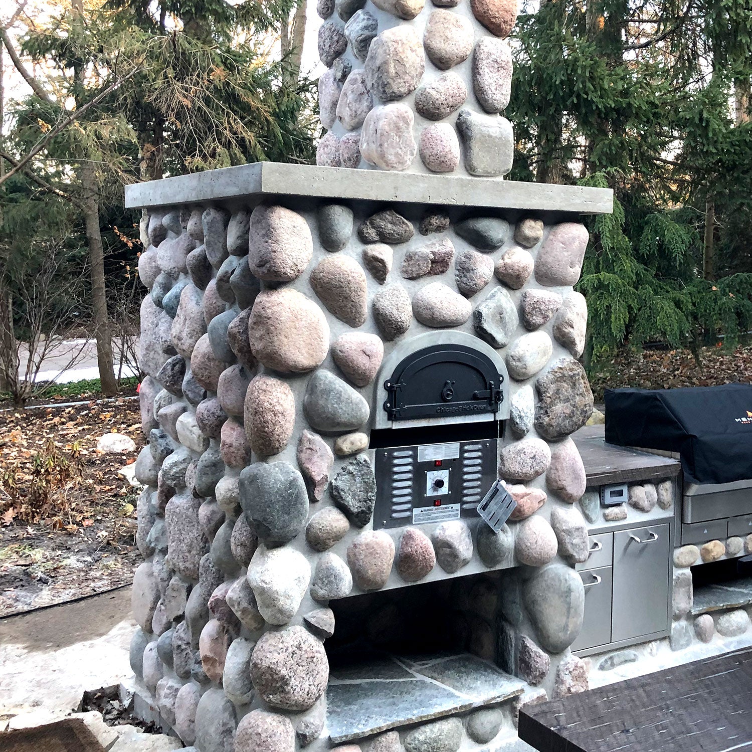 The CBO 750 Hybrid DIY Kit (Residential) from Chicago Brick Oven is a rustic, dual-fuel (gas and wood) outdoor pizza oven, featuring large, round stones and a central metal door. As part of an integrated outdoor kitchen setup surrounded by greenery and trees, it includes a top chimney and side metal storage cabinets reinforced with refractory cement.