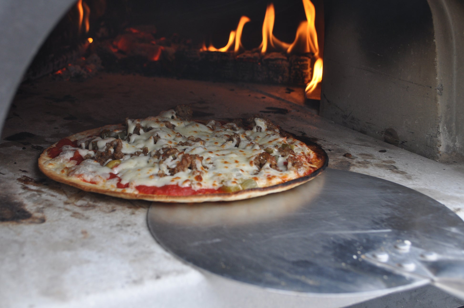 A pizza is being taken out of a wood-fired oven with the Chicago Brick Oven Aluminum Pizza Peel - Turning Pizza Paddle. The pizza, topped with melted cheese and various toppings, is slightly browned around the edges. The flames and burning wood in the background emit a warm glow, highlighting the precision of this essential baking tool from Chicago Brick Oven, featuring a leather strap and detachable wood handle.