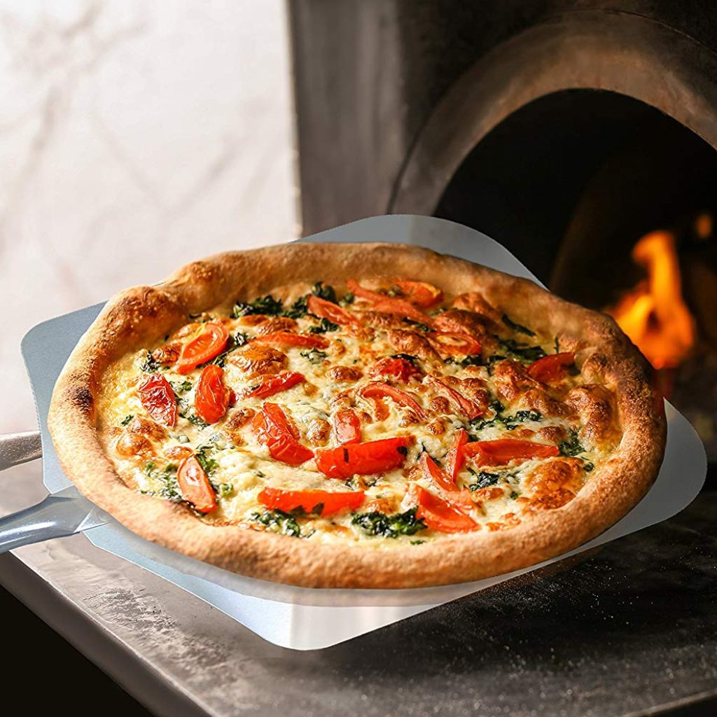 Using the Chicago Brick Oven Pizza Peel 12" x 14" - a 35.5" long aluminum metal pizza paddle with a detachable 21" wooden handle from Chicago Brick Oven, a freshly baked pizza topped with tomato slices, spinach, and melted cheese is being removed from a traditional wood-fired oven. The pizza features a golden-brown crust and is set against the background of the oven's orange glow.