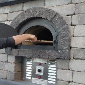 A person places a wooden pizza peel with dough into the CBO 750 Hybrid DIY Kit (Residential) by Chicago Brick Oven. This high-quality outdoor pizza oven, known for its stone facade and metal control panel with various dials and buttons, supports both gas and wood as fuel options. The person's arm is visible, extending into the oven.