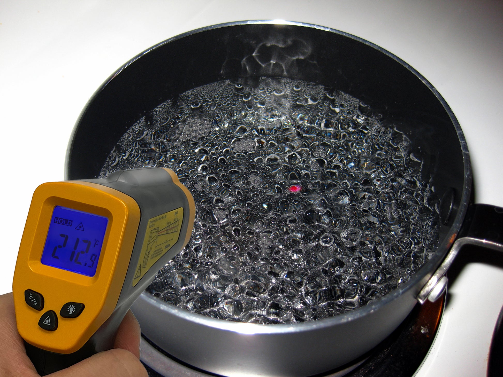 A close-up of a stainless steel pot filled with boiling water on a stove. A hand is holding a CBO Home Infrared Thermometer Gun aimed at the water's surface, with the thermometer's screen displaying a reading of 21.2°C.