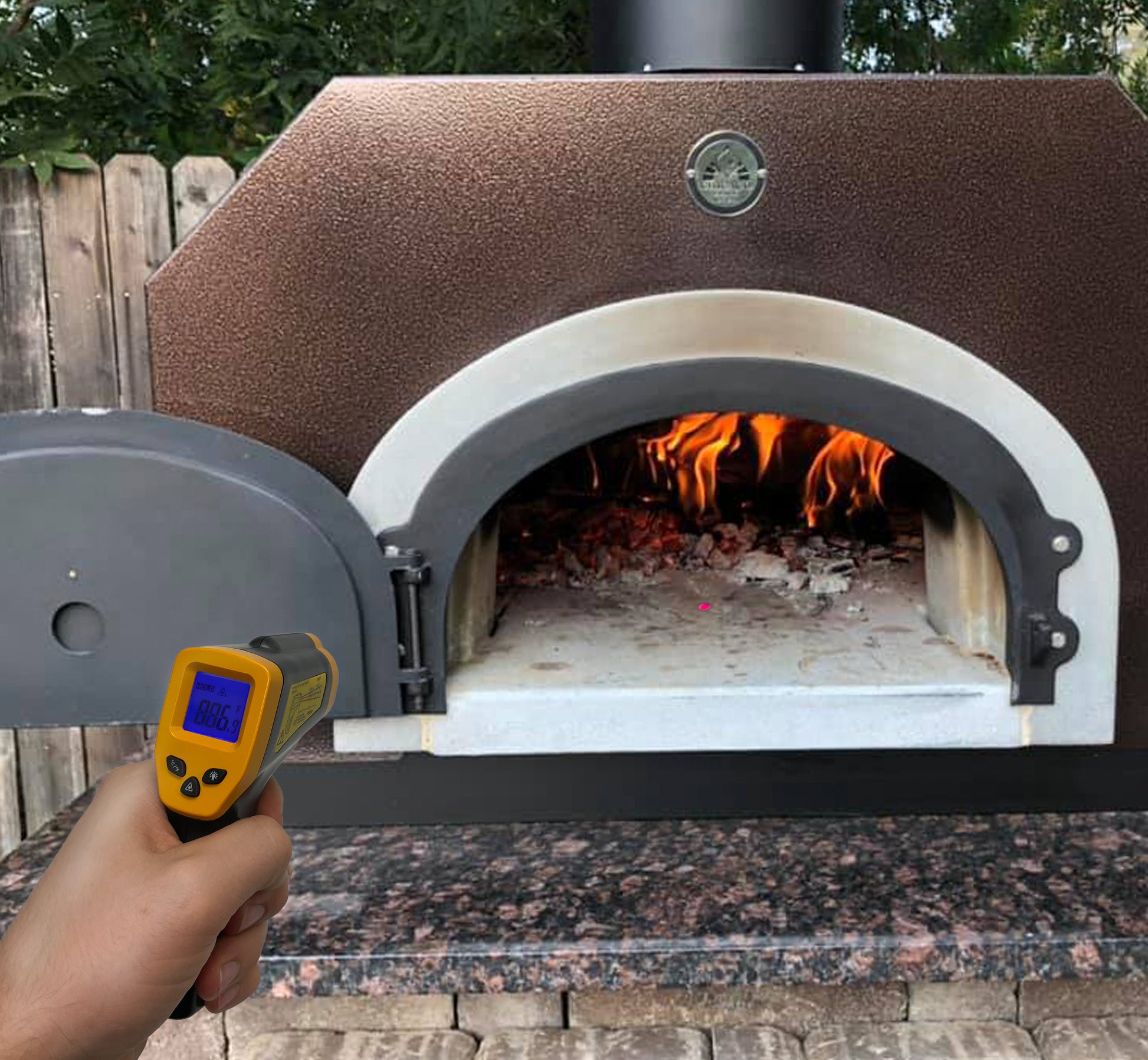 A person holds a CBO Home Infrared Thermometer Gun in front of a wood-fired oven with an open door. The digital thermometer displays a reading of 886°F as burning wood and embers glow inside. This essential pizza oven accessory from CBO Home, suitable for temperatures ranging from -58°F to 1022°F, sits on a stone surface surrounded by bricks.