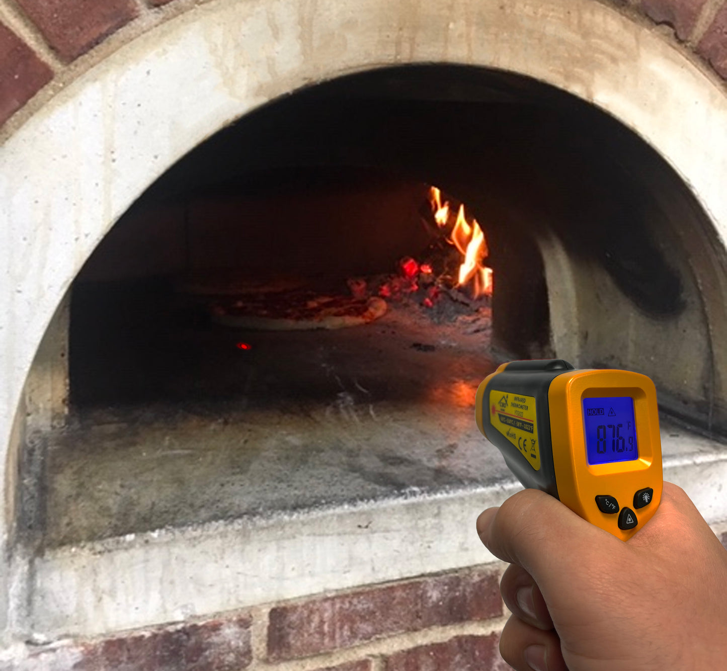 A hand holds the CBO Home Infrared Thermometer Gun, a vital pizza oven accessory, in front of a brick pizza oven, measuring the temperature at 876 degrees Fahrenheit. Inside the oven, a fire burns and a pizza is being cooked on the stone surface. The thermometer's wide temperature range display is illuminated.