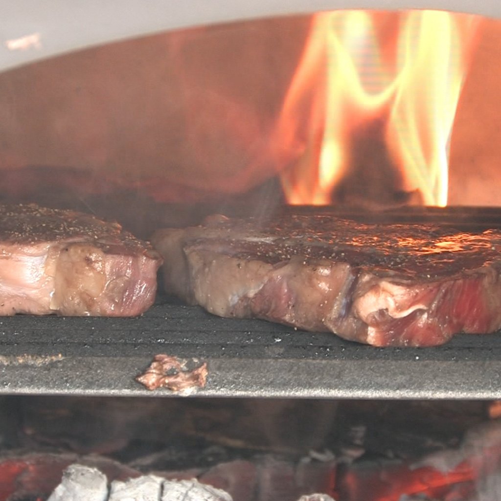 Two thick cuts of steak sizzle on a charcoal grill inside the Chicago Brick Oven CBO 750 Hybrid Stand (Commercial) | Dual Fuel (Gas and Wood), with bright flames roaring in the background. The edges of the meat are slightly charred, indicating they are in the process of being grilled to perfection, just like they would be in an outdoor pizza oven.