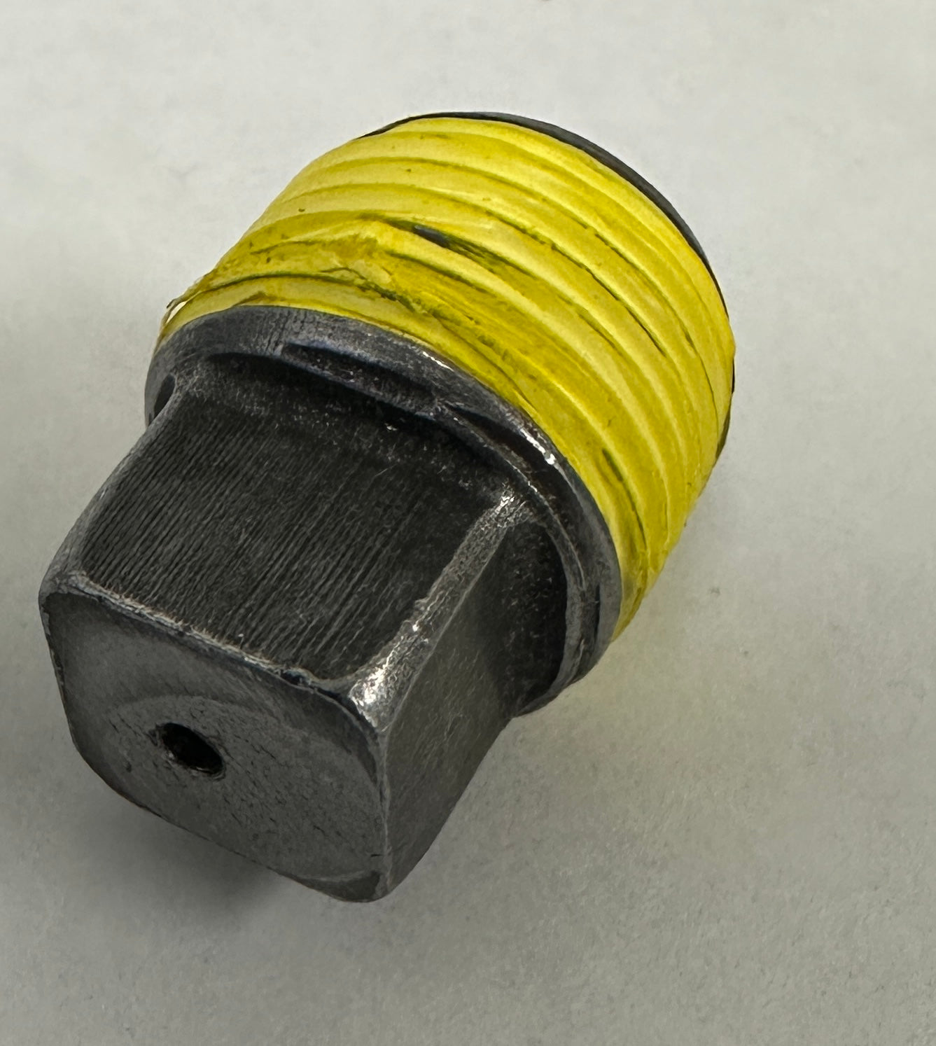 An image of the Hybrid Americano LP Orifice from Chicago Brick Oven, a metal bolt featuring a square-shaped head and threads wrapped in yellow Teflon tape, resembling an LP orifice. The bolt is positioned diagonally against a plain, light-colored background.