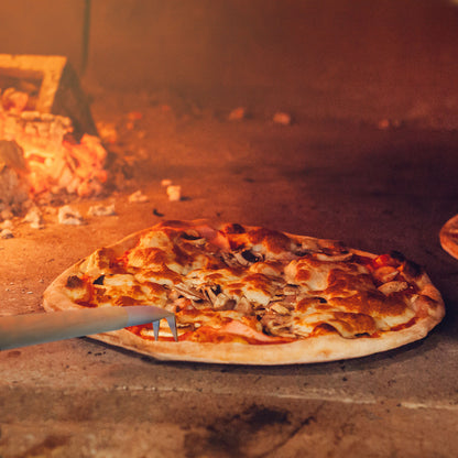 A freshly baked pizza with bubbling cheese and toppings is being expertly maneuvered out of a quality pizza oven using the Chicago Brick Oven's Aluminum 17.5 inch Pizza Spinner Turning Fork with a Wooden Handle and Leather Strap. The rustic, stone interior glows with the heat of burning embers in the background, ensuring safety and perfect cooking.